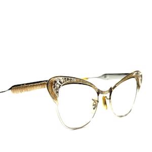 Op de afbeelding: Vintage cat-eye bril met een goudkleurig montuur. De bovenste randen zijn versierd met heldere en rode strass-steentjes. De veren hebben een getextureerde gouden afwerking en de oortips zijn lichtbruin.