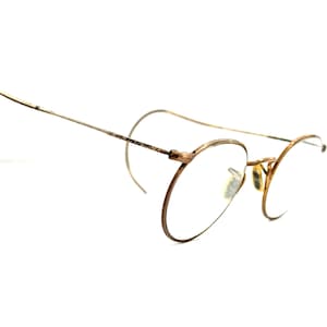 May include: Vintage gold-tone eyeglasses with round lenses and curved temples. The glasses have a delicate wire frame and are set against a white background. The design is classic and elegant, suggesting a retro or antique style.