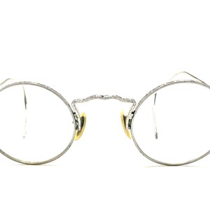 May include: Silver-toned, round-framed eyeglasses with clear lenses. The frames feature intricate detailing along the top and sides, with small nose pads. The arms are thin and extend to the temples. The glasses are set against a white background.