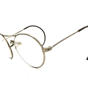 Puede incluir: Gafas vintage con montura de metal plateado y lentes transparentes. Las gafas tienen una forma redonda clásica y patillas curvas con terminales negros. La montura está detallada con un diseño texturizado. Las gafas están sobre un fondo blanco.
