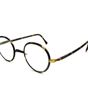 May include: Round eyeglasses with a black frame and tortoiseshell arms. The lenses are clear, and the glasses have a gold-coloured bridge. The glasses are set against a white background.