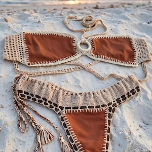 Può includere: Un bikini a due pezzi con pannelli in tessuto marrone tinta unita bordati da rifiniture in uncinetto color crema. Il top include un anello centrale in legno e laccetti da annodare, mentre lo slip presenta una vita in uncinetto con volant.