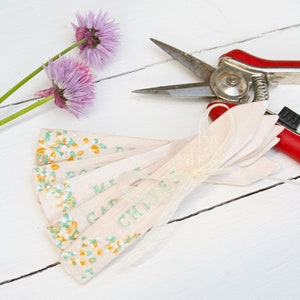 May include: Pale pink garden markers with floral patterns and text, tied with a sheer ribbon. A pair of red-handled garden shears and sprigs of purple chive blossoms are also present, all on a white wooden surface.