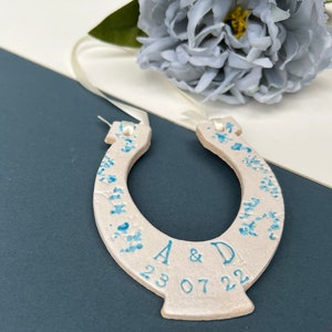 White ceramic glazed horseshoe with pretty blue shades of flowers up the sides, with text: INITIAL & INITIAL DATE. Text and date along the bottom. Cream ribbon attached to the top for hanging purposes.