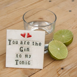 May include: A white ceramic coaster with the text "You are the Gin to my Tonic" in black letters. Two red hearts are at the top of the coaster. A glass of water and two lime wedges are on a wooden surface.