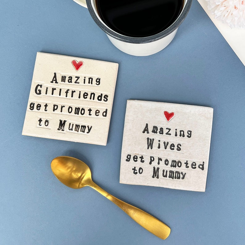 Two square ceramic coasters finished in an off white glaze. Black text reads, amazing wives get promoted to mummy or amazing girlfriends get promoted to mummy. Small red heart above the text. Gold teaspoon.