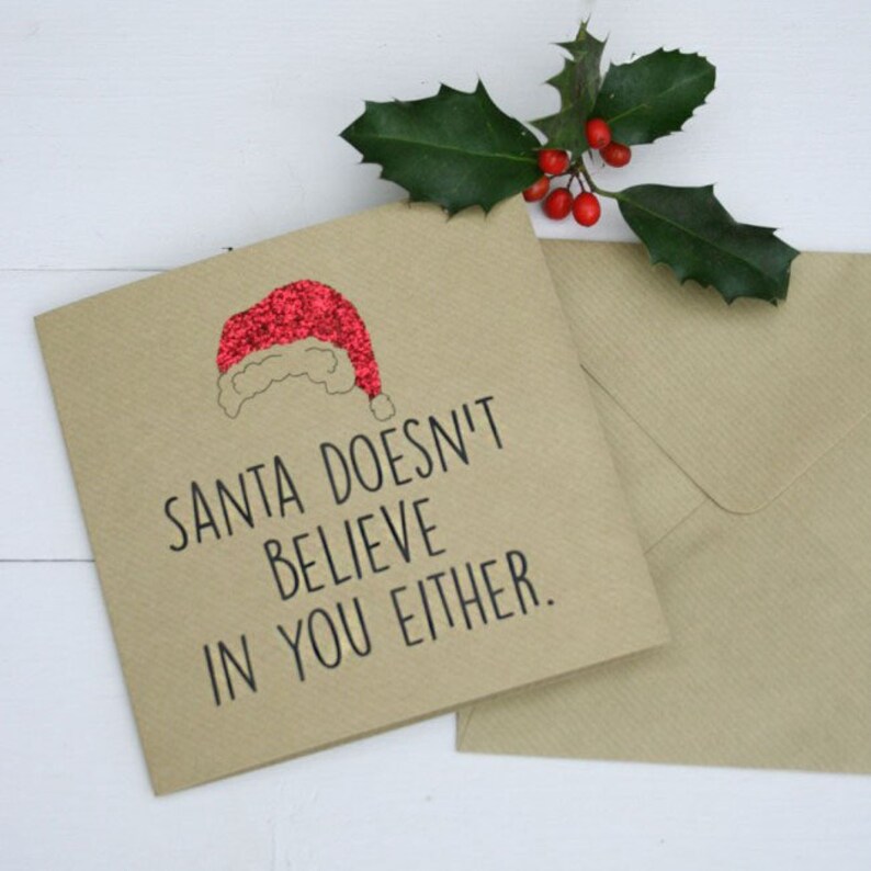 Square craft card with black text: SANTA DOESN'T BELIEVE IN YOU EITHER. 
A red glittery Santa hat sits at the top of the card.