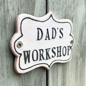 May include: White metal sign with black lettering that reads "Dad's Workshop". The sign has a decorative border and is attached to a wooden surface with screws.