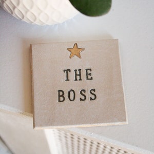 May include: A beige ceramic coaster with a gold star and the text "THE BOSS" in black.