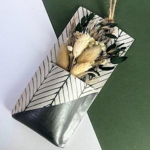 May include: A wall-mounted pocket planter featuring a white surface with black hand-painted chevron patterns and a solid dark grey base. The pocket contains a dried floral arrangement with beige bunny tails, white baby's breath, and dark green leaves.