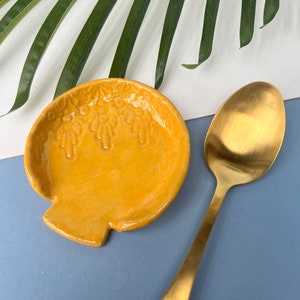 May include: A yellow ceramic spoon rest with a decorative design. The spoon rest is shaped like a shell and has a smooth, glossy finish.