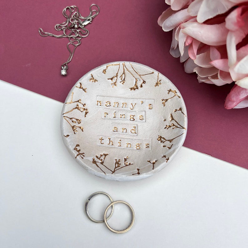 Approx 9cm round white glaze dish. Text: nanny's rings and things. Written in gold, as are the flowers imprinted around the edge of the dish.