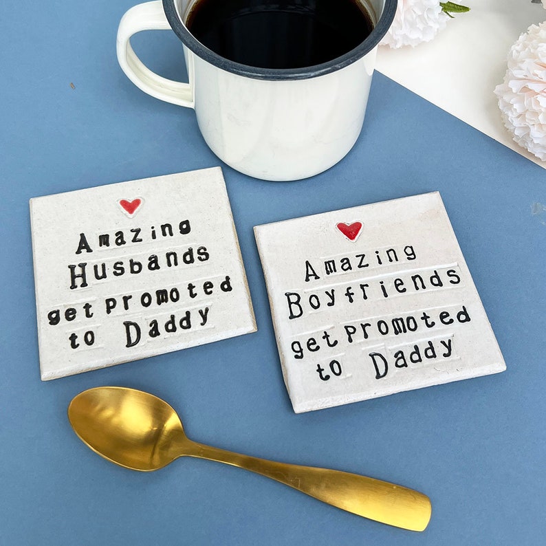 Two square ceramic coasters finished in an off white glaze. Black text reads, amazing husbands get promoted to daddy or amazing boyfriends get promoted to daddy. Small red heart above the text. Gold teaspoon.