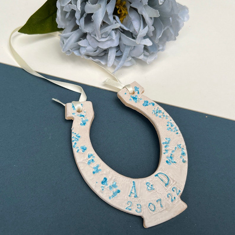 White ceramic glazed horseshoe with pretty blue shades of flowers up the sides, with text: INITIAL & INITIAL DATE. Text and date along the bottom. Cream ribbon attached to the top for hanging purposes.