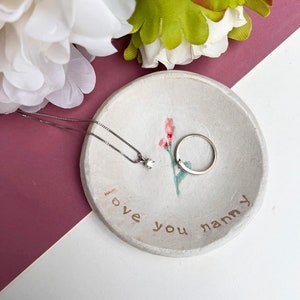 May include: White ceramic trinket dish with a floral design and the words "love you nanny" written on it. A silver necklace with a small diamond pendant and a silver ring are sitting in the dish.