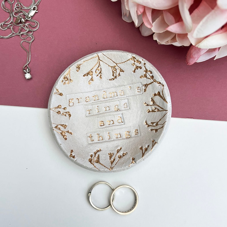 Approx 9cm round white glaze dish. Text: grandma's rings and things. Written in gold, as are the flowers imprinted around the edge of the dish.