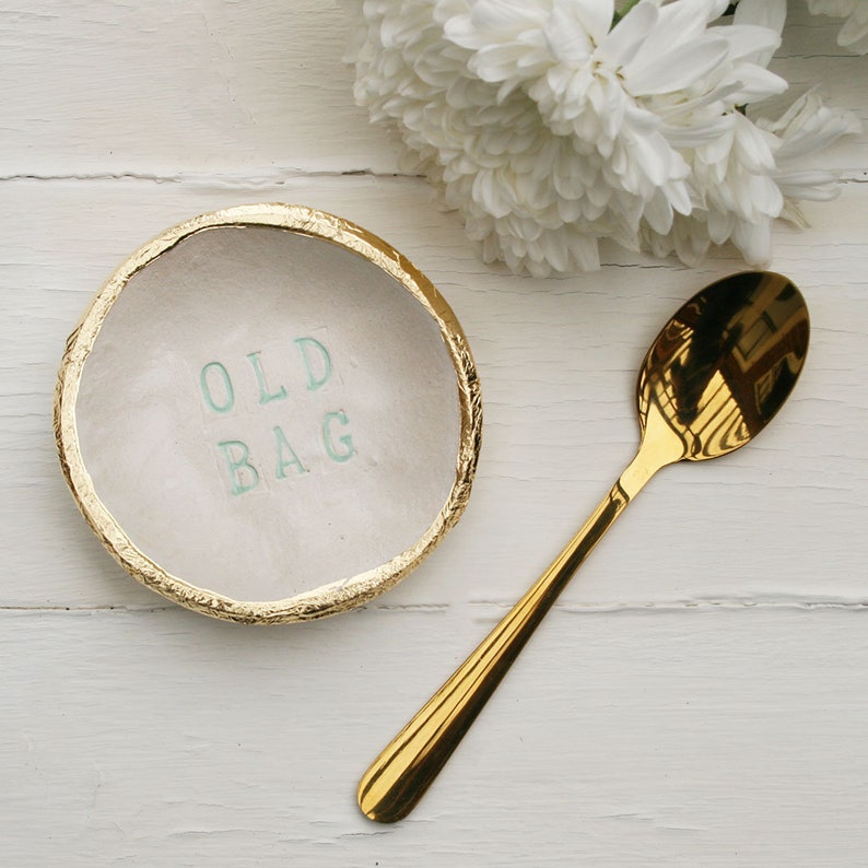 Small round ceramic dish, finished in off white glaze. The text old bag printed in the centre and hand painted in sage green. Gold leaf applied to the rim of the dish. Gold teaspoon.