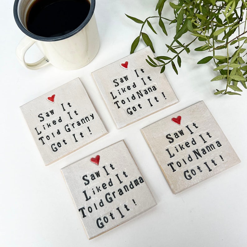 K&ouml;nnte beinhalten: Set aus vier wei&szlig;en Keramik-Untersetzern mit roten Herzen und schwarzem Text. Jeder Untersetzer tr&auml;gt die Aufschrift "Saw It Liked It Told [Name] Got It!" wobei der Name Granny, Nana, Grandma und Nanna ist.