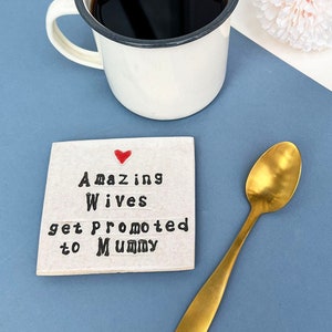 Square ceramic coaster finished in an off white glaze. Black text reads, amazing wives get promoted to mummy. Small red heart above the text. Gold teaspoon.