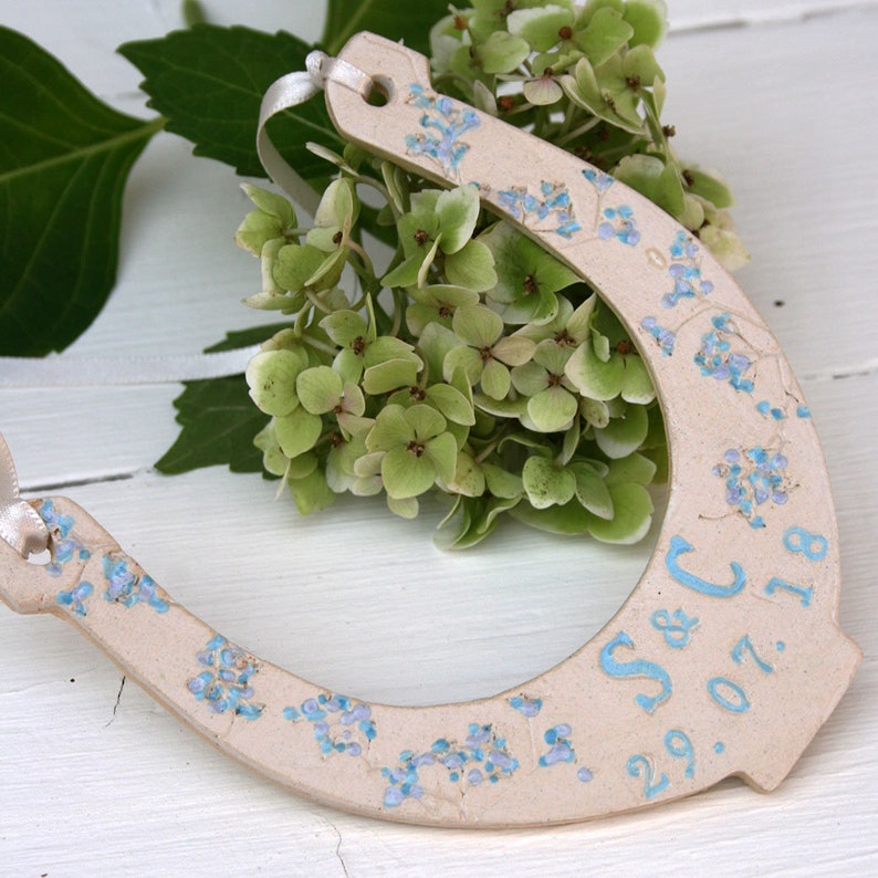 White ceramic glazed horseshoe with pretty blue shades of flowers up the sides, with text: INITIAL & INITIAL DATE. Text and date along the bottom. Cream ribbon attached to the top for hanging purposes.