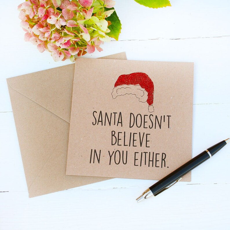 Square craft card with black text: SANTA DOESN'T BELIEVE IN YOU EITHER. 
A red glittery Santa hat sits at the top of the card.