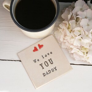 white square coaster with two hearts at the top with the same black text: We love you daddy