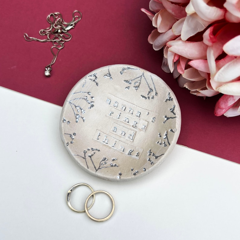 Approx 9cm round white glaze dish. Text: nanna's rings and things. Written in silver, as are the flowers imprinted around the edge of the dish.