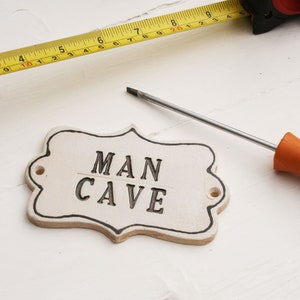 A off white ceramic plaque sign with black capital writing MAN CAVE, black lined edging with 2 screw holes