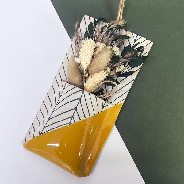 May include: A wall-mounted pocket vase featuring a white and black chevron pattern on the upper section and a solid mustard yellow glazed finish on the lower portion, containing a dried floral arrangement of beige grasses and dark green leaves.