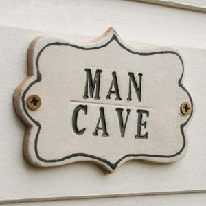 A off white ceramic plaque sign with black capital writing MAN CAVE, black lined edging with 2 screw holes