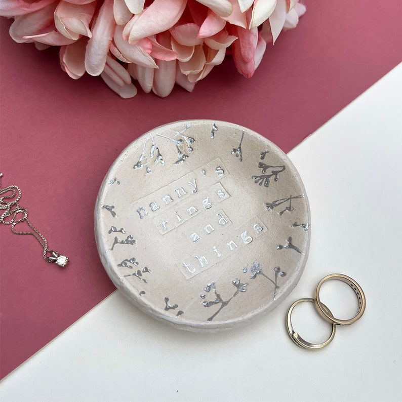 Approx 9cm round white glaze dish. Text: nanny's rings and things. Written in silver, as are the flowers imprinted around the edge of the dish.