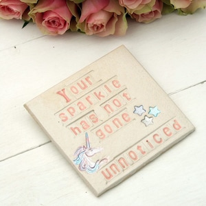 May include: A light brown ceramic tile with the words "Your sparkle has not gone unnoticed" in pink letters. The tile has a unicorn and three stars on it.