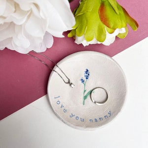 May include: White ceramic trinket dish with a blue floral design and the text "love you nanny". A silver necklace with a small diamond pendant and a silver ring are on the dish.