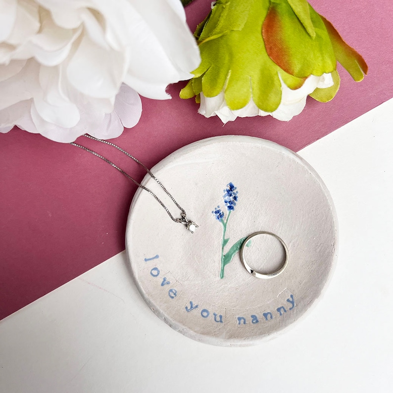 May include: White ceramic trinket dish with a blue floral design and the text "love you nanny". A silver necklace with a small diamond pendant and a silver ring are on the dish.