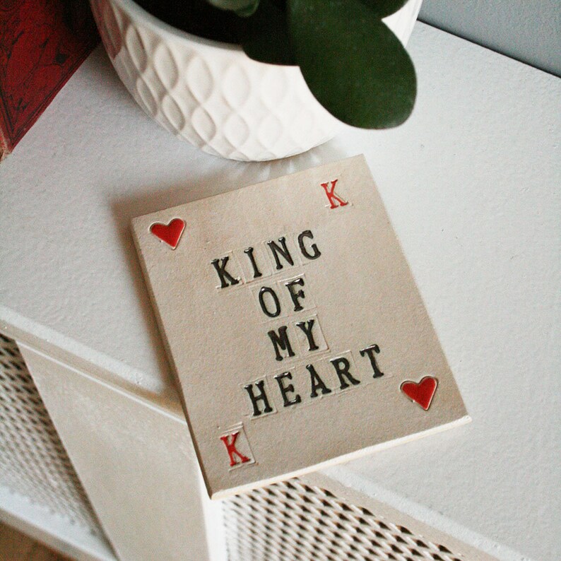 small white ceramic rectangle coaster with black text king of my heart
in left top corner and bottom right corners are red hearts and red k in the other corners