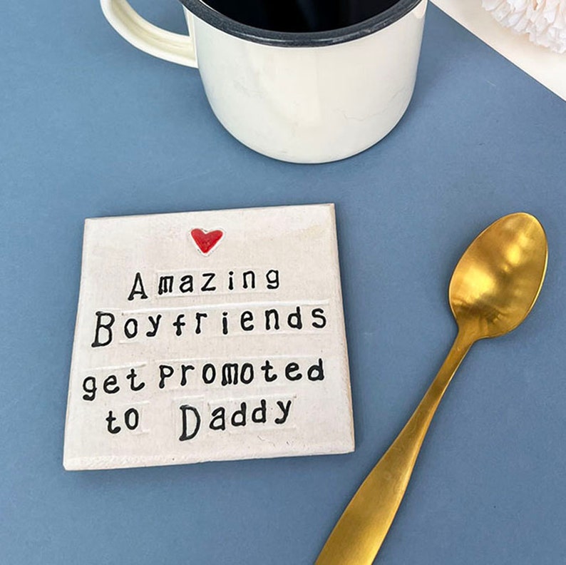 Square ceramic coaster finished in an off white glaze. Black text reads, amazing boyfriends get promoted to daddy. small red heart above the text. Gold teaspoon.