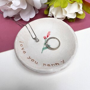 May include: White ceramic trinket dish with a floral design and the words "I love you nanny" written in brown paint. A silver ring and necklace are sitting in the dish.