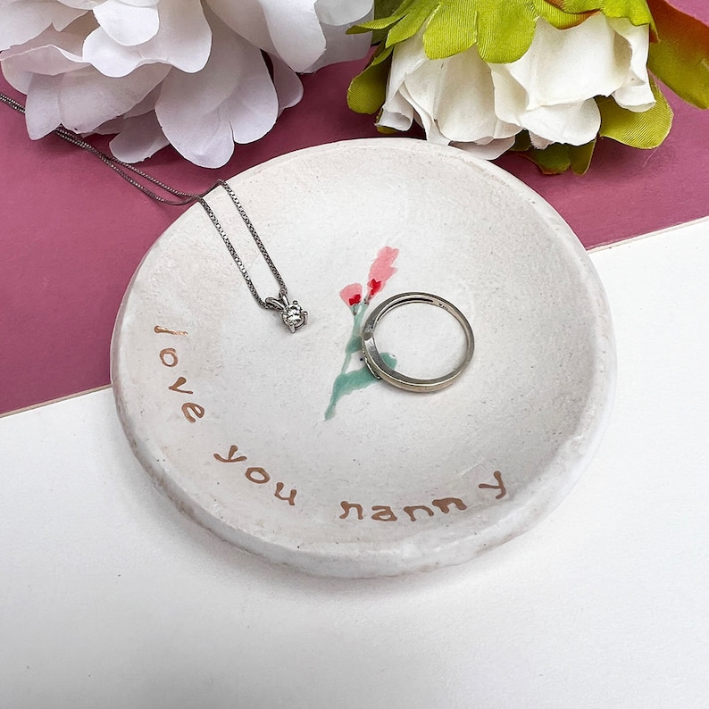 May include: White ceramic trinket dish with a floral design and the words "I love you nanny" written in brown paint. A silver ring and necklace are sitting in the dish.