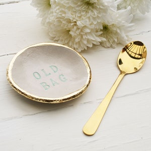 Small round ceramic dish, finished in off white glaze. The text old bag printed in the centre and hand painted in sage green. Gold leaf applied to the rim of the dish. Gold teaspoon.