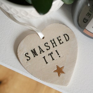 May include: A white heart-shaped ceramic ornament with the words "SMASHED IT!" in black letters and a gold star.