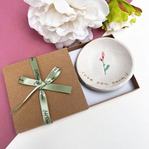 May include: A small white ceramic dish with a pink flower design and the words "I love you nanny" in gold lettering. The dish is in a brown gift box with a green ribbon.
