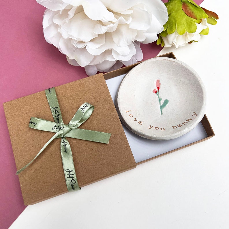 May include: A small white ceramic dish with a pink flower design and the words "I love you nanny" in gold lettering. The dish is in a brown gift box with a green ribbon.