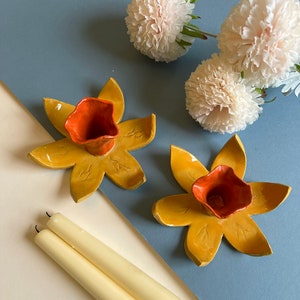 May include: Two ceramic candle holders shaped like daffodils with orange centres. The candle holders are on a blue surface with white flowers.