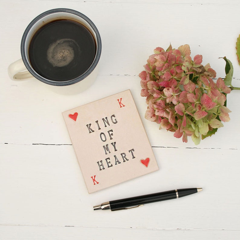 small white ceramic rectangle coaster with black text king of my heart
in left top corner and bottom right corners are red hearts and red k in the other corners