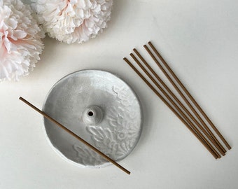 May include: A white ceramic incense burner with a floral design and a small hole in the center. Six brown incense sticks are laid out next to the burner.