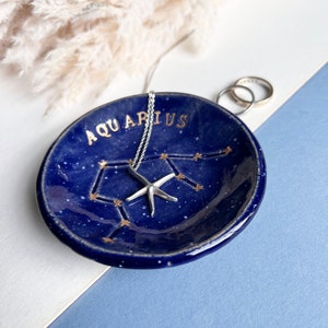 Könnte beinhalten: Eine blaue Keramikschale mit goldenen Sternen und dem Wort "Aquarius" in goldenen Buchstaben. Eine silberne Halskette mit einem Seestern-Anhänger liegt in der Schale.