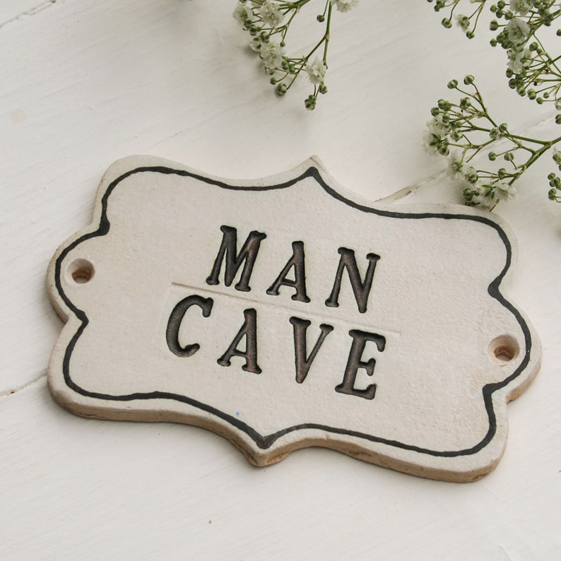 A off white ceramic plaque sign with black capital writing MAN CAVE, black lined edging with 2 screw holes