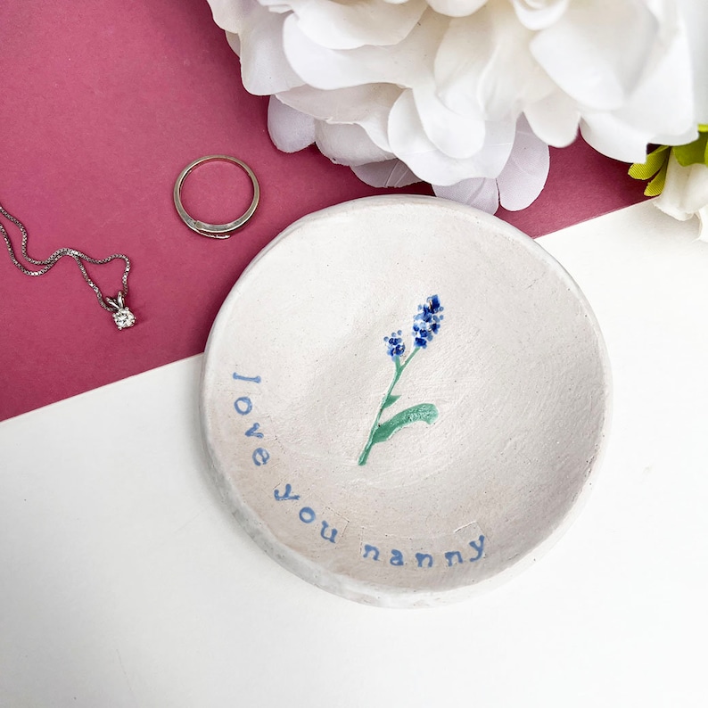 May include: White ceramic ring dish with a blue flower design and the text "I love you nanny" written in blue.