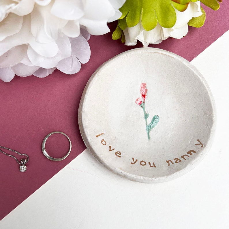 May include: White ceramic ring dish with a pink flower design and the words "Love you nanny" written in gold.