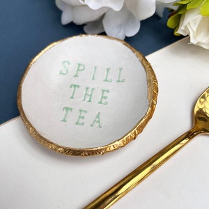 May include: White ceramic ring dish with a gold rim. The dish has the words "Spill the Tea" written in green.